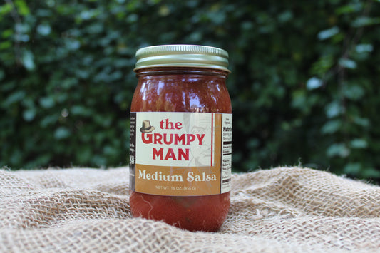 A jar of 'The Grumpy Man' Medium Salsa sits on a textured burlap cloth with a blurred green garden background.
