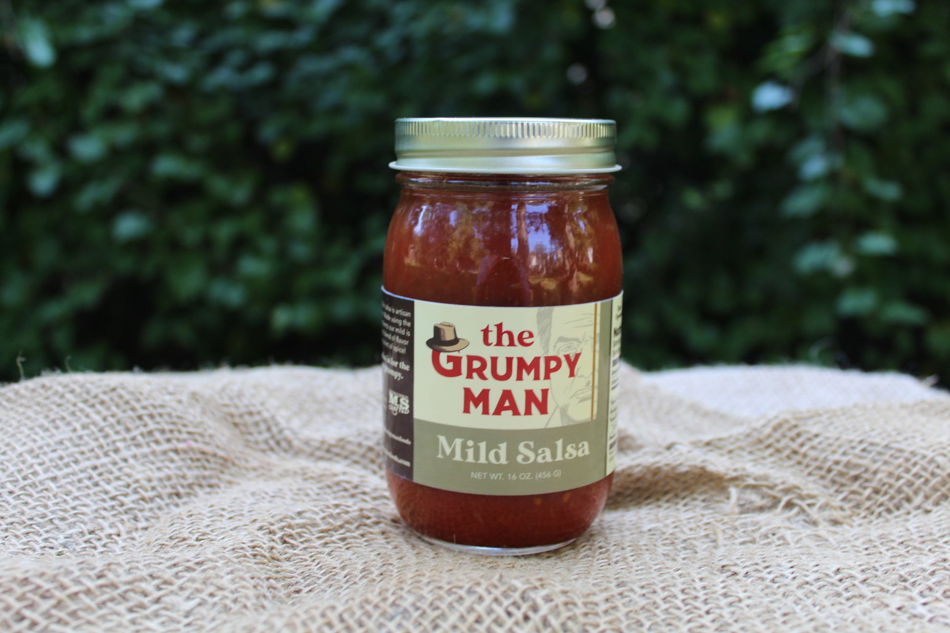 A glass jar of Mild Salsa by The Grumpy Man sits on a textured burlap cloth with a blurred green garden background.