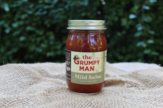 A glass jar of Mild Salsa by The Grumpy Man sits on a textured burlap cloth with a blurred green garden background.