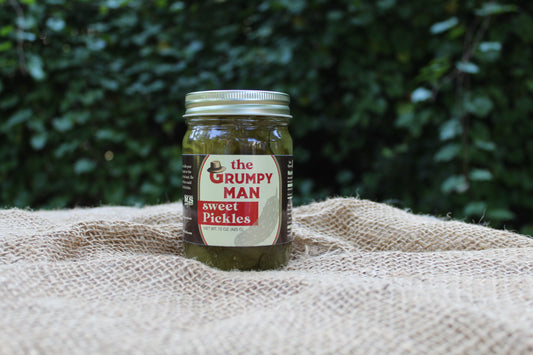 A jar of 'The Grumpy Man' Sweet Pickles sits on a textured burlap cloth with a blurred background of the farm behind them.